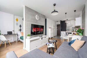 Una sala de estar moderna con un sofá gris, un televisor mostrando 'NETFLIX', un escritorio con una computadora portátil y una zona de comedor al fondo.