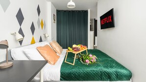Ett välordnat hotellrum med en säng, ett sängbord med en lampa, en väggmonterad TV och ett bord med frukt och blommor.