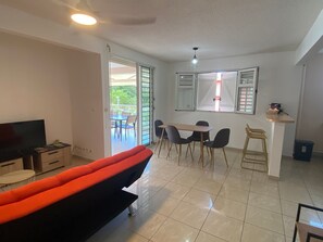 A living room with a sofa, TV, dining table, chairs, and a balcony with outdoor furniture. - 2-Bedroom Apartment - Walk to Beach & Pool (Les Trois-Ilets)