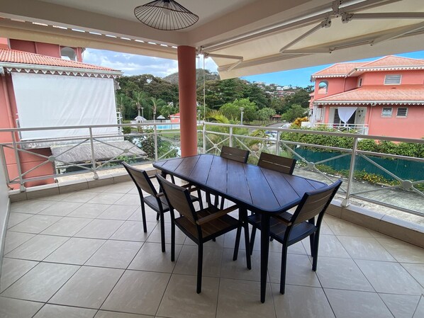 A covered outdoor area with a table and chairs, a tiled floor, and a view of buildings and greenery.