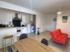A compact living space with a kitchenette, a dining area, a red sofa, and a washing machine. - 2-Bedroom Apartment - Walk to Beach & Pool (Les Trois-Ilets)
