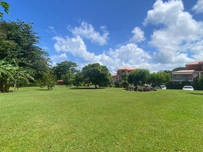 A grassy area with trees and a building with a red roof. - 2-Bedroom Apartment - Walk to Beach & Pool (Les Trois-Ilets)