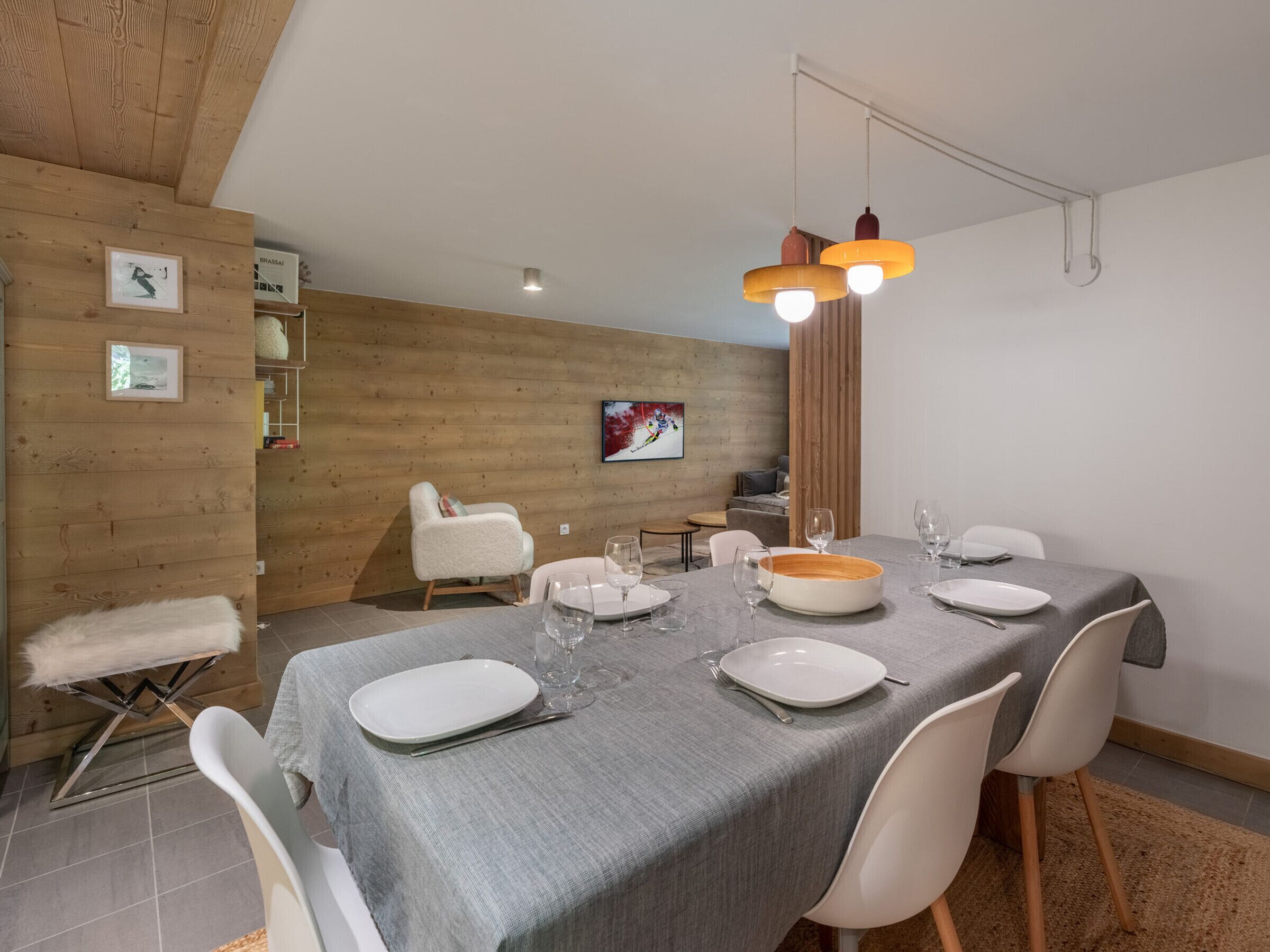 A dining area with a wooden wall, a white chair, a dining table set with plates and glasses, and a hanging light fixture.