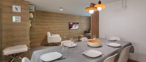 A dining area with a wooden wall, a white chair, a dining table set with plates and glasses, and a hanging light fixture.