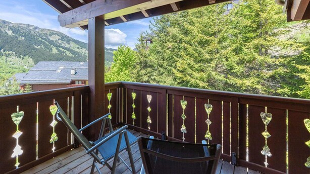A wooden deck with two chairs and a table, overlooking a mountainous landscape with trees and a building in the background.