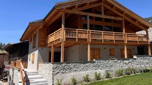 A two-story wooden chalet with a balcony, stone wall, and a staircase leading to the entrance.