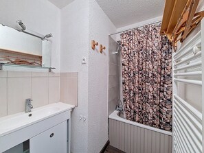 Ein Badezimmer mit einem weißen Waschbecken, einem Spiegel, einer Dusche mit einer Duschvorhang aus Kieselsteinen und einem Holzregal über dem Spiegel.