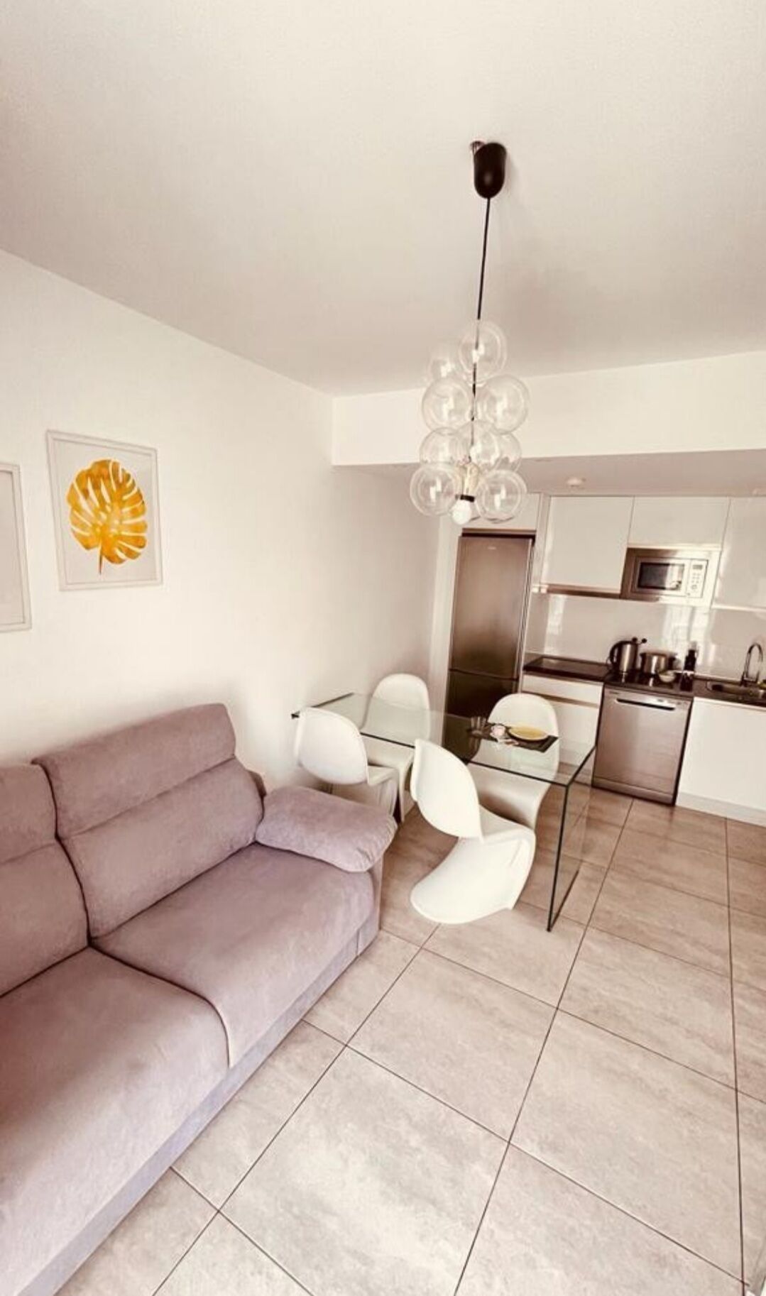 A modern living room with a grey sofa, a dining table with chairs, a kitchen area with a microwave, and a unique spherical chandelier.