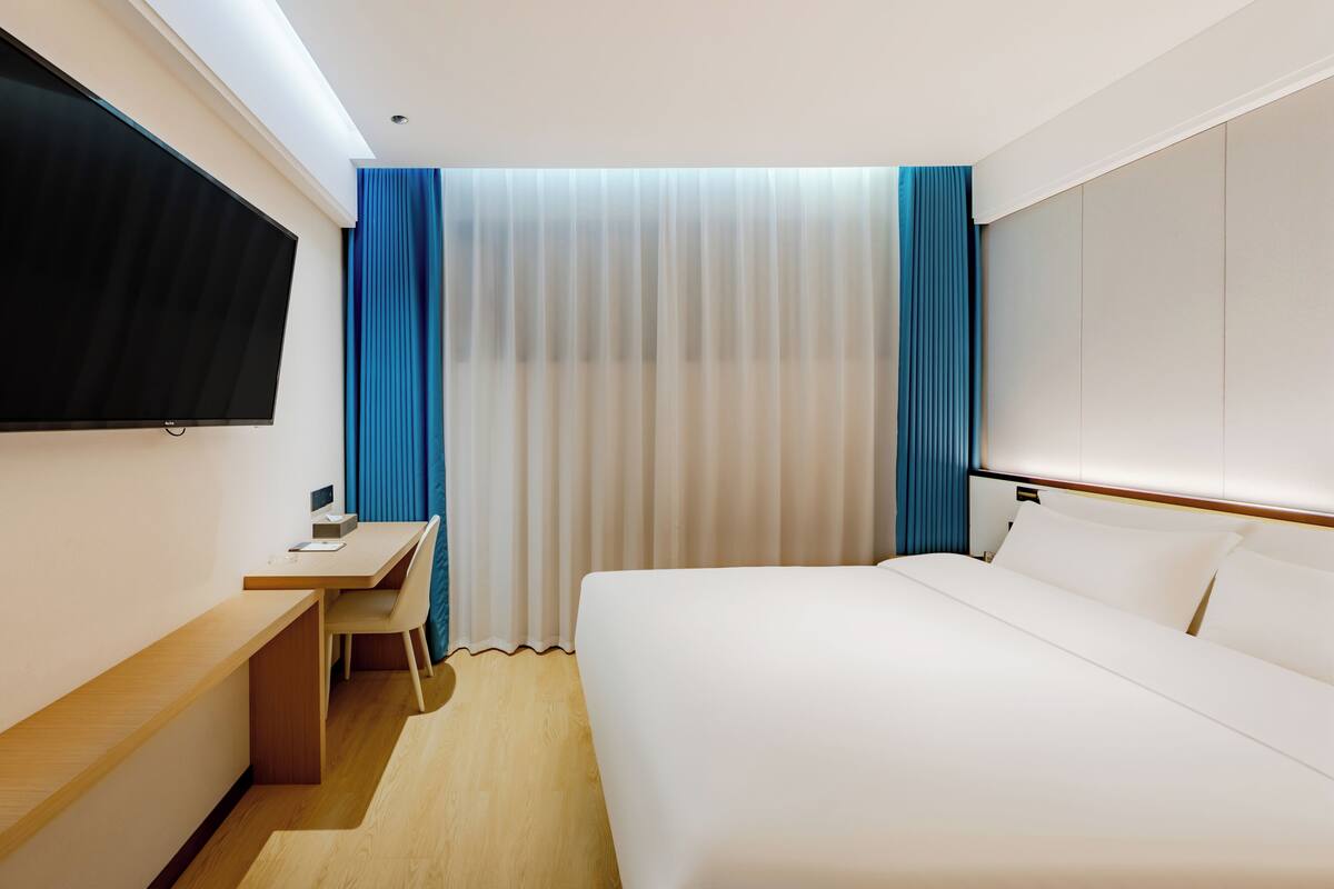 A hotel room with a bed, a desk, a chair, a flat-screen TV, and curtains.