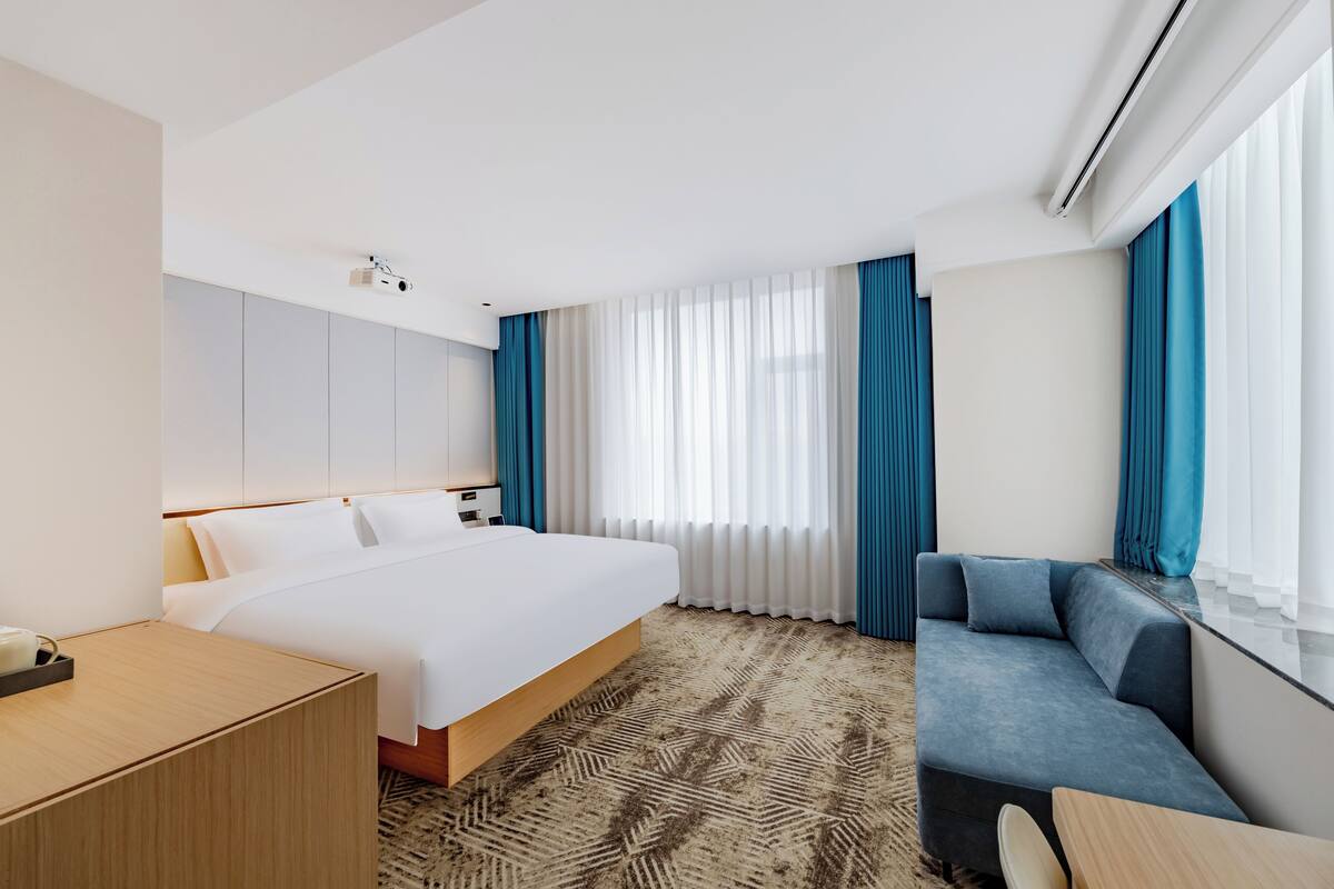 A modern hotel room with a large bed, a desk, a sofa, and a large window with curtains.