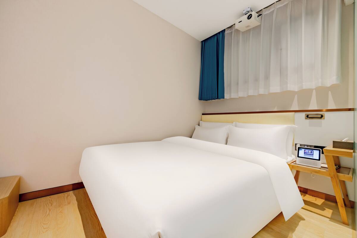 A hotel room with a large bed, a bedside table with a lamp, a small desk with a computer, and a window with curtains.
