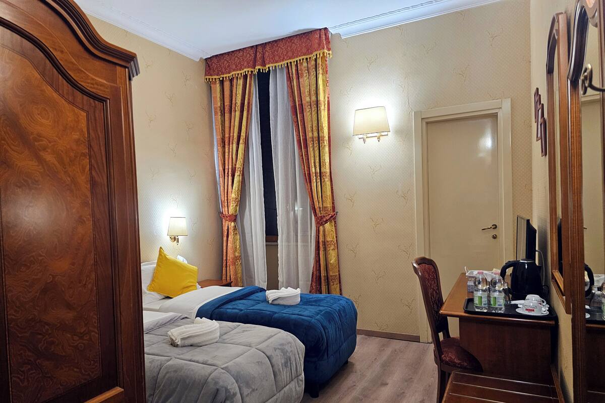 A hotel room with two beds, a wooden wardrobe, a desk with toiletries, and a mirror.