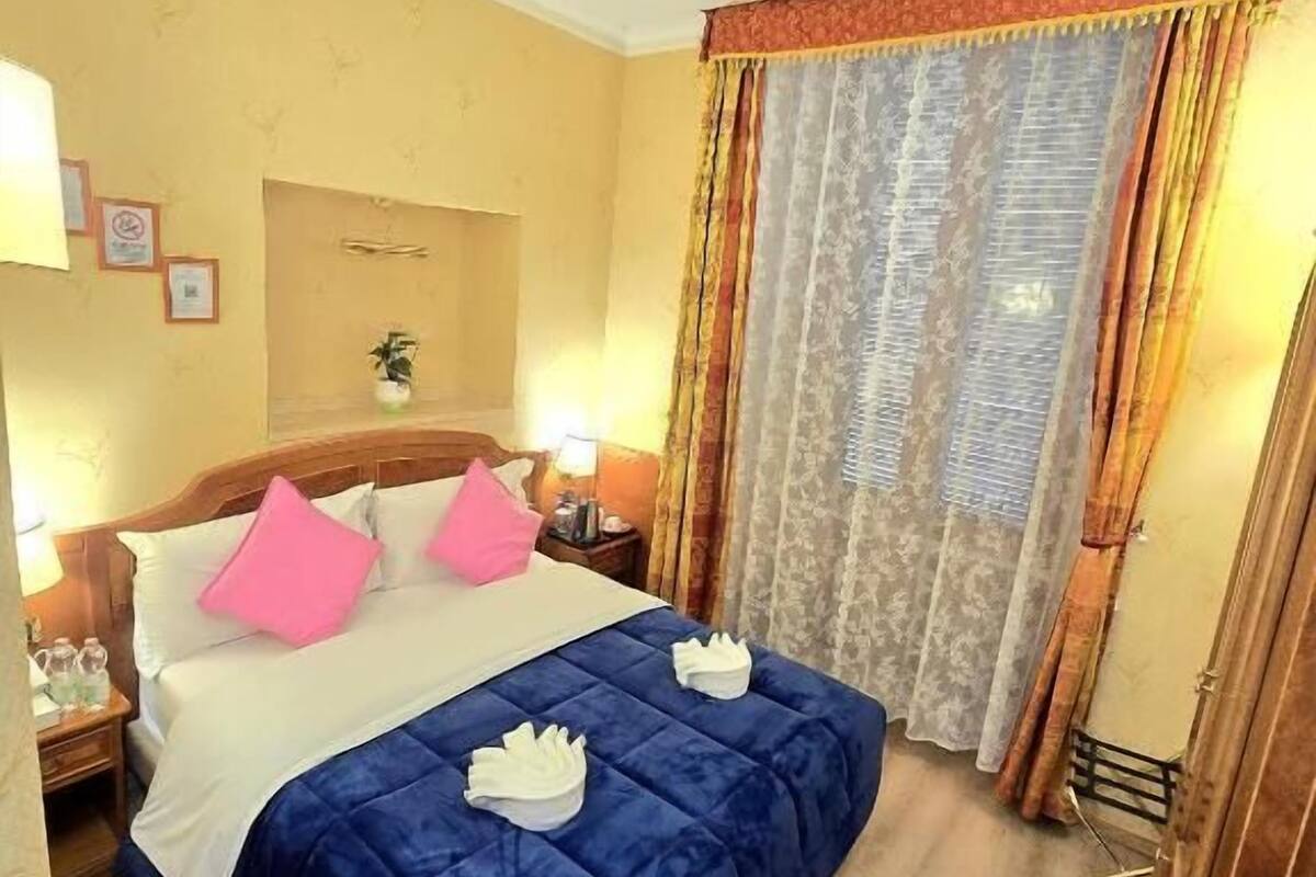 A hotel room with a bed, pillows, a nightstand, a lamp, and a window with curtains.