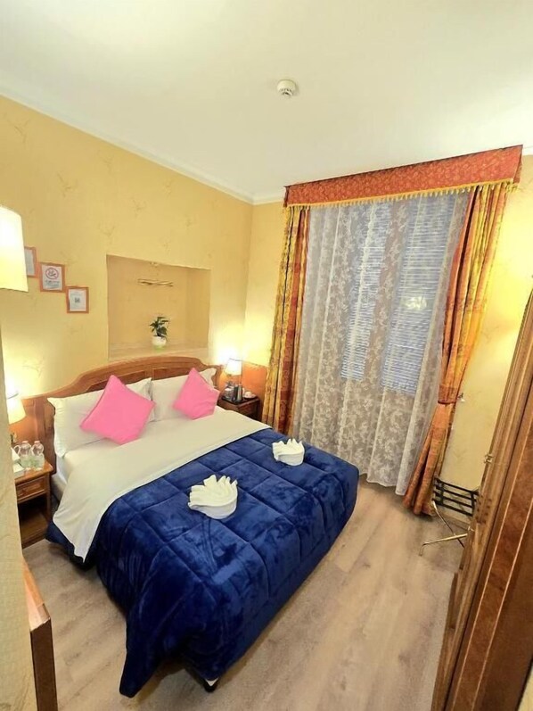 A hotel room with a bed, pillows, a nightstand, a lamp, and a window with curtains. - Hotel Amadeus (Rome)