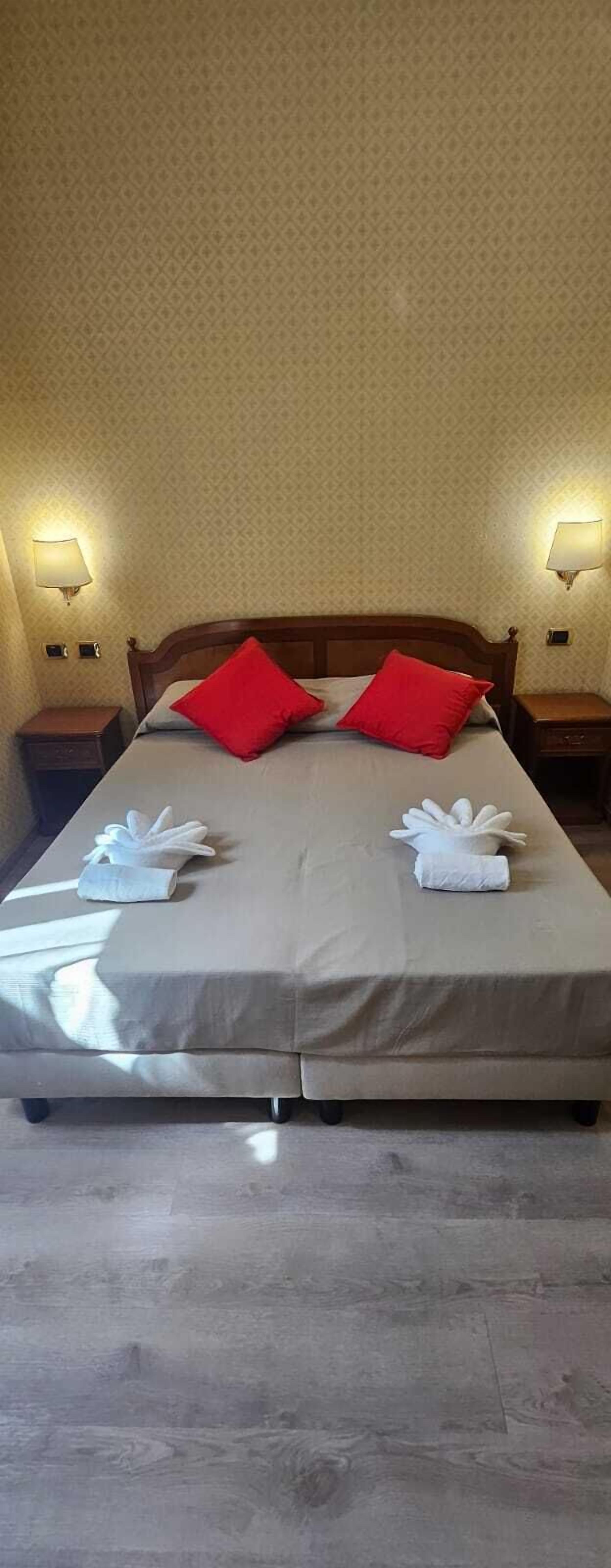 A neatly made bed with red pillows, a white towel, and a white robe on the bed. There are two bedside tables with lamps on either side of the bed.