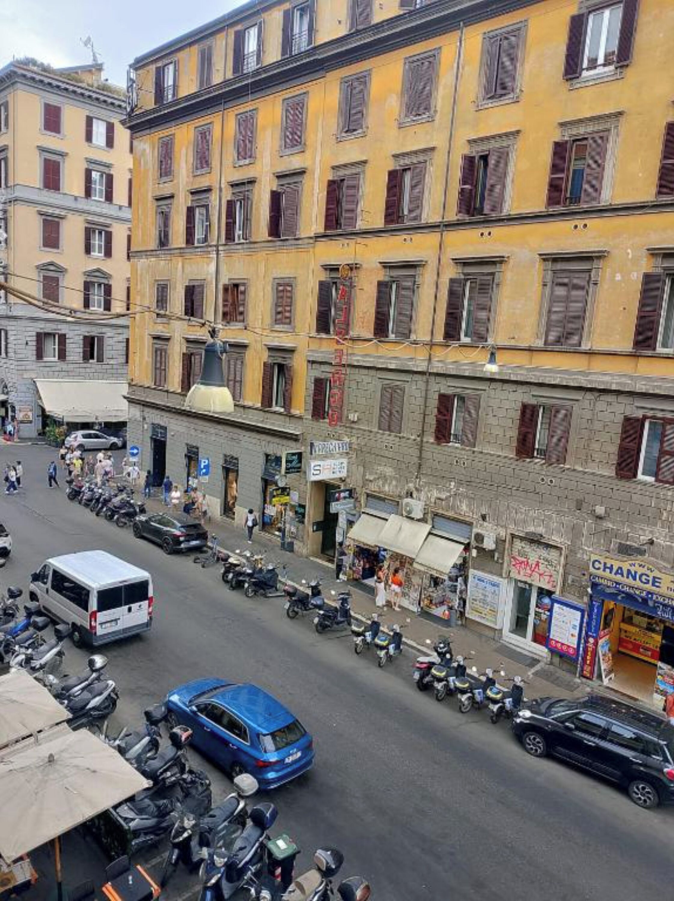 A street view with a row of yellow buildings, parked vehicles, and pedestrians.