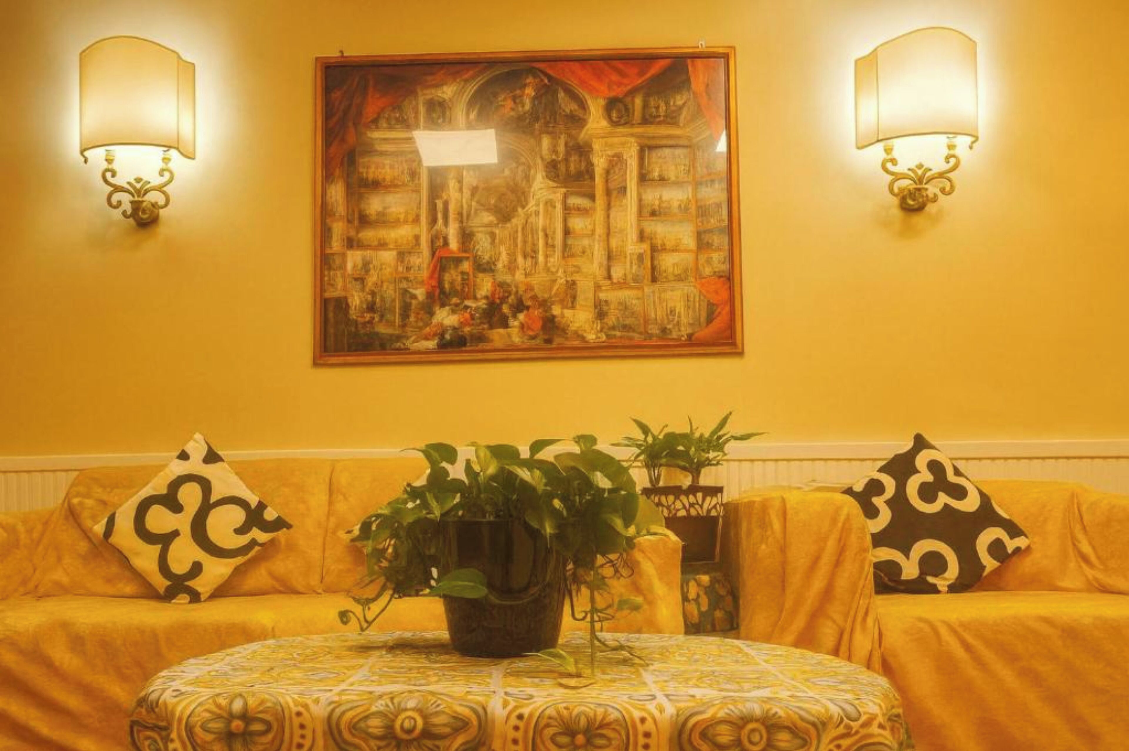 A room with a yellow sofa, two wall-mounted lamps, a framed painting, and a table with a patterned tablecloth.