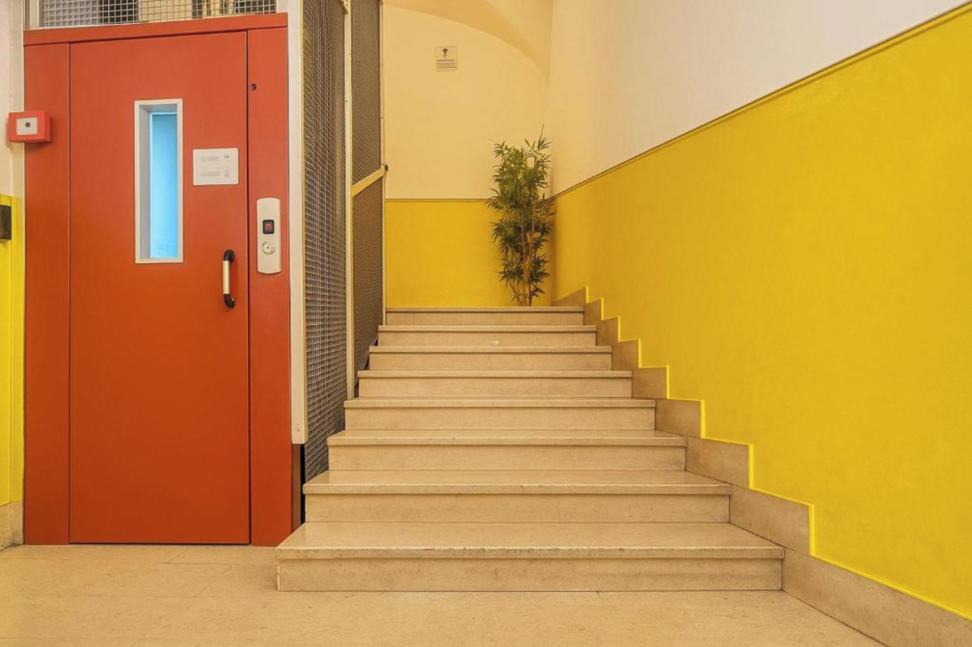 A staircase with beige steps leading up to a landing, flanked by yellow walls and a red door with a light fixture.