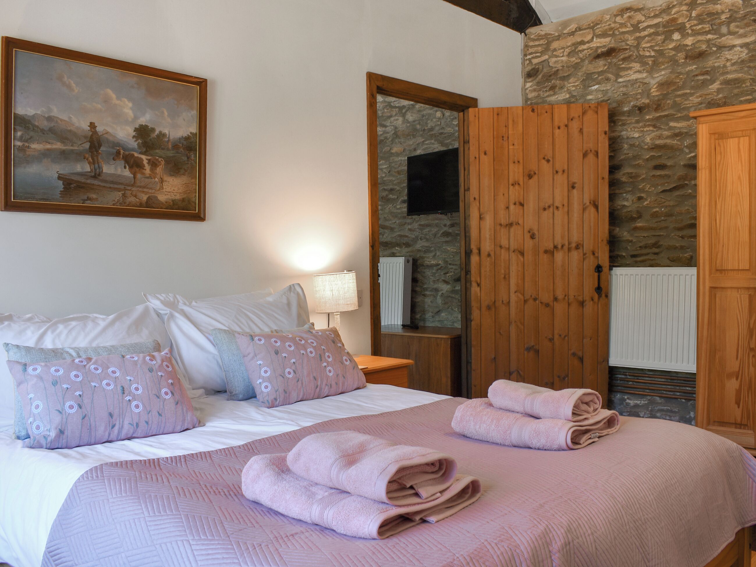 A bedroom with a bed, pillows, a painting, a wooden wardrobe, and a stone wall.
