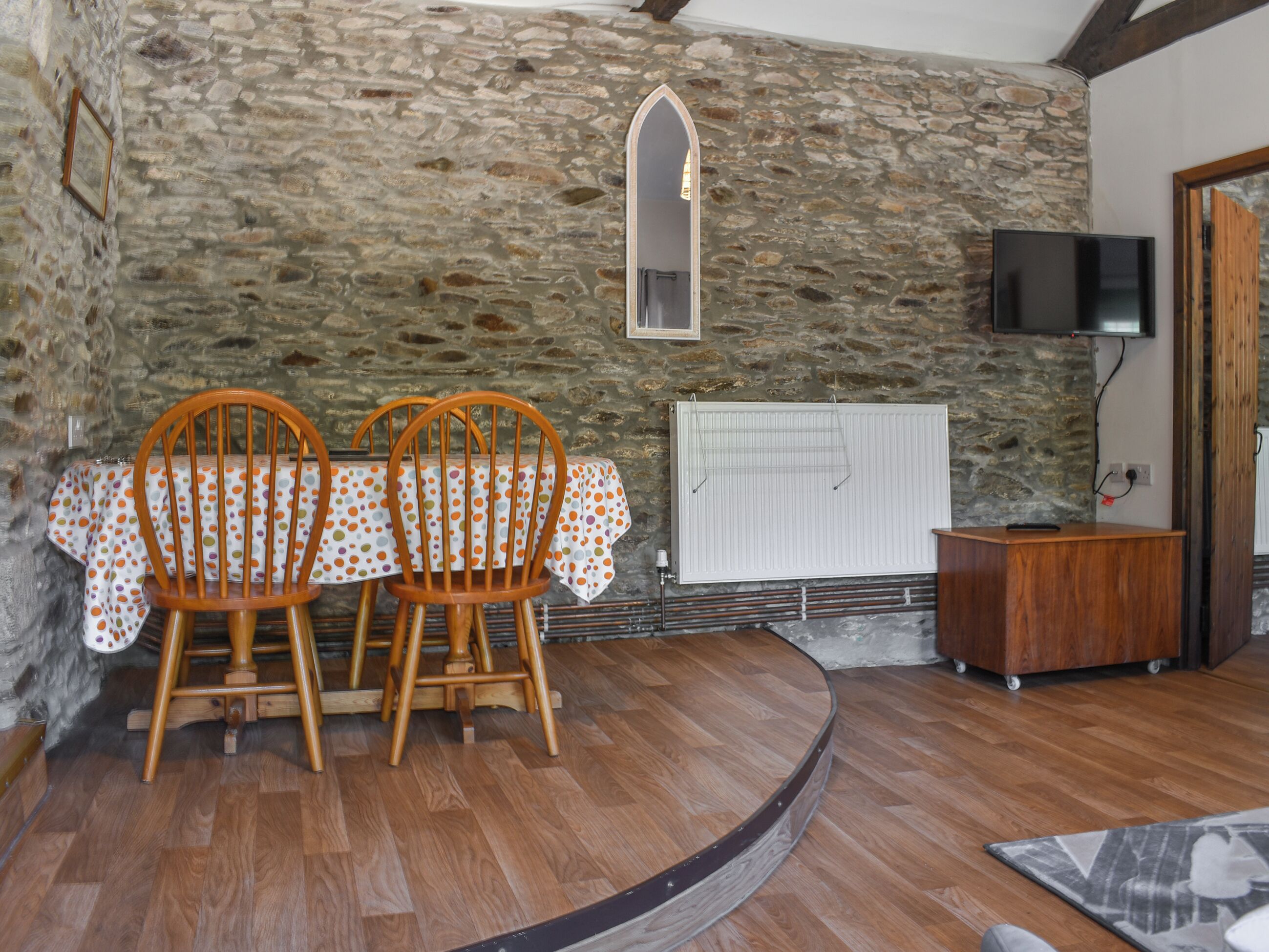 A room with a stone wall, a wooden dining table with chairs, a radiator, a television, and a wooden cabinet.