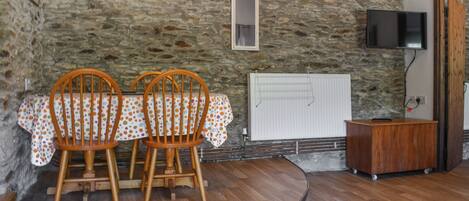 A room with a stone wall, a wooden dining table with chairs, a radiator, a television, and a wooden cabinet.