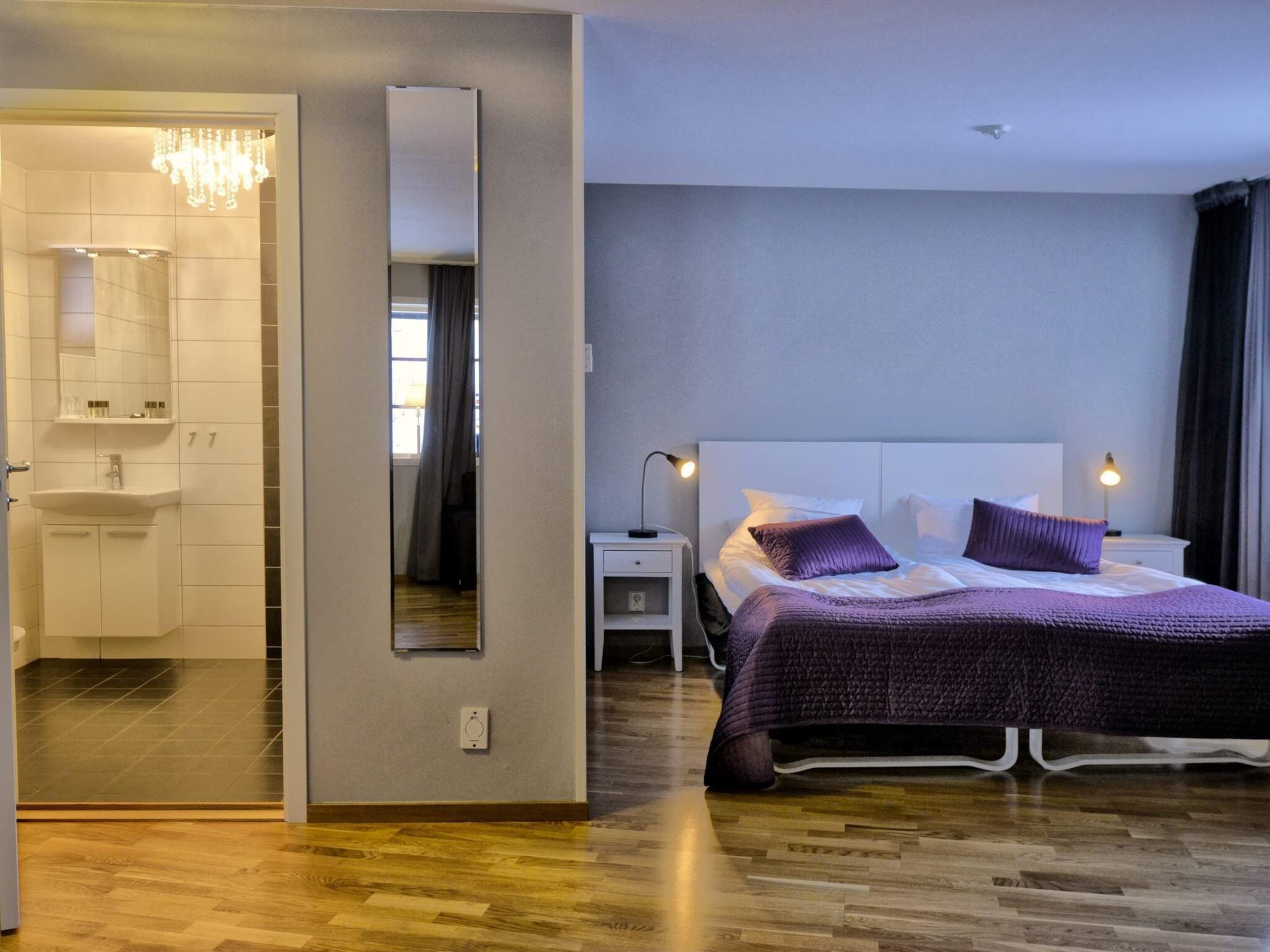 A modern hotel room with a large bed, bedside lamps, a mirror, and a bathroom with a sink and shower.