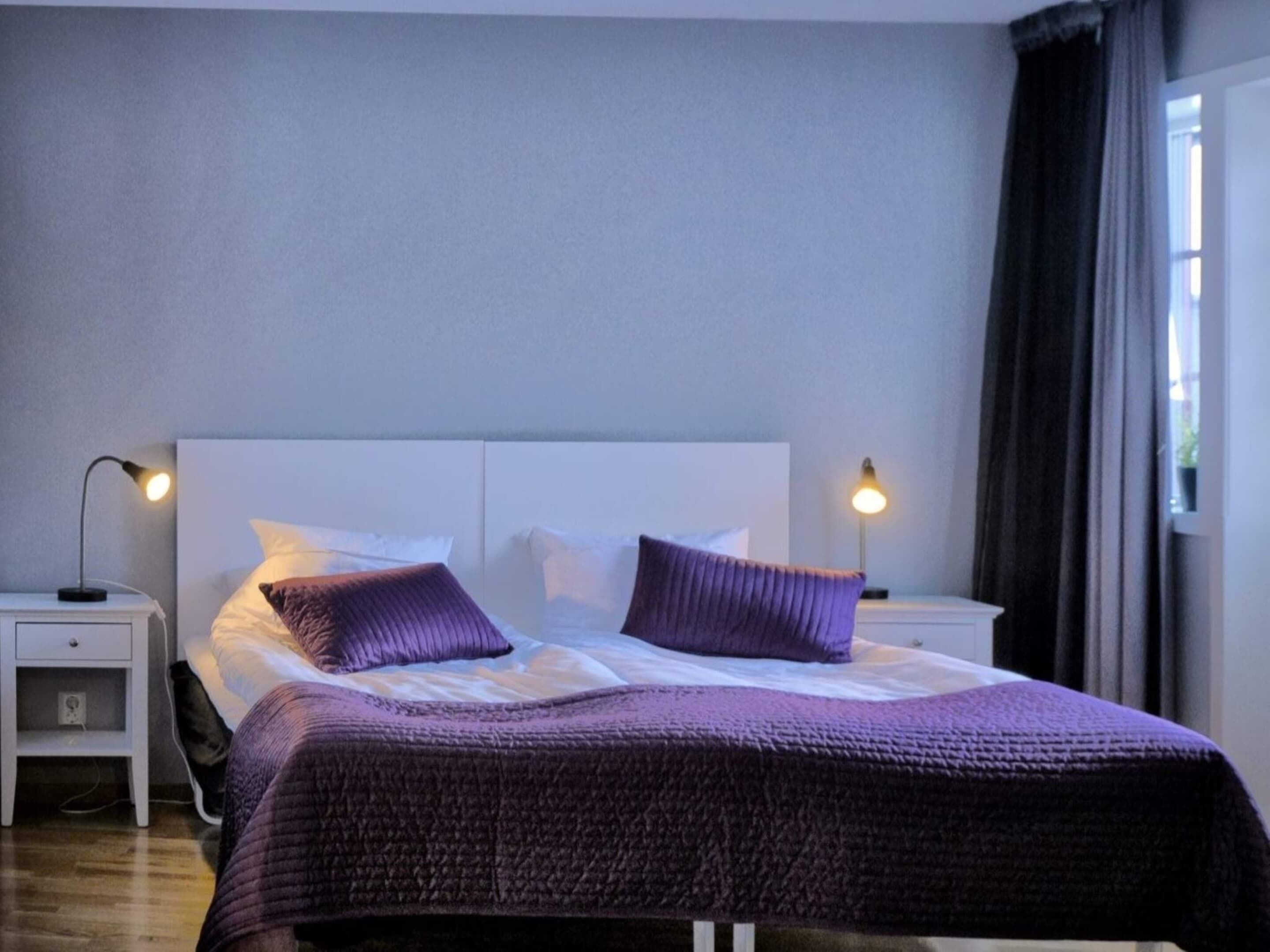 A neatly made bed with a headboard, two bedside lamps, and a window with curtains.