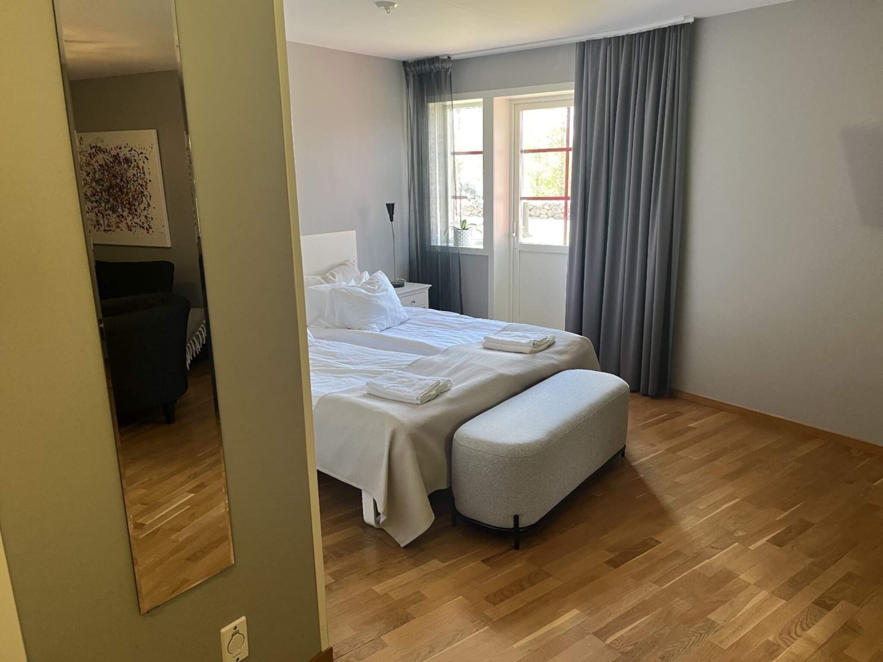 A bedroom with a bed, a bedside table, a chair, a window with curtains, and a wooden floor.