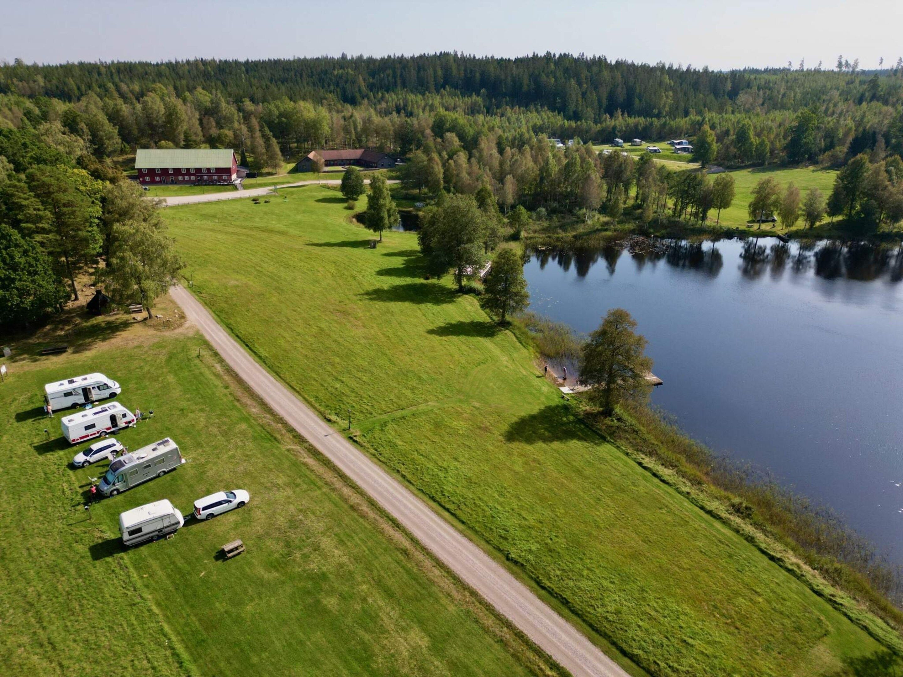 An aerial view of a campsite with RVs parked along a road, a lake, and forested hills in the background.