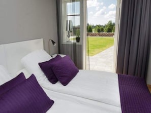Une chambre à coucher avec un lit, des oreillers, une table de chevet, une lampe et une vue sur un jardin, visible à travers une porte vitrée.