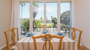 Un comedor con una mesa preparada para cuatro personas, ventanas amplias con vistas a palmeras y edificios, y sillas de madera con mantel a rayas.