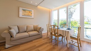 Una sala de estar con un sofá color beige, una mesa de comedor de madera y sillas. Se ve una ventana con vistas a palmeras y edificios.