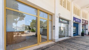 Entrada de un edificio con grandes puertas de vidrio, un banco de madera afuera y tiendas, incluyendo 'Lisgardente' y 'Zometime'.