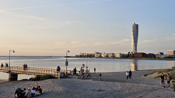 A riverside promenade with people walking, sitting, and cycling. A distinctive tower-like building is visible in the background. - 5 Person Holiday Home in Stehag (Stehag)