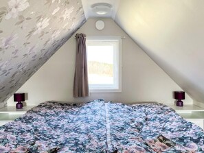 Un dormitorio con techo inclinado, una ventana con cortinas, una cama con colcha estampada y dos lámparas de noche.