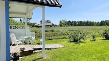 A covered outdoor seating area with a table and chairs, a wooden deck, and a view of a green field and distant buildings.