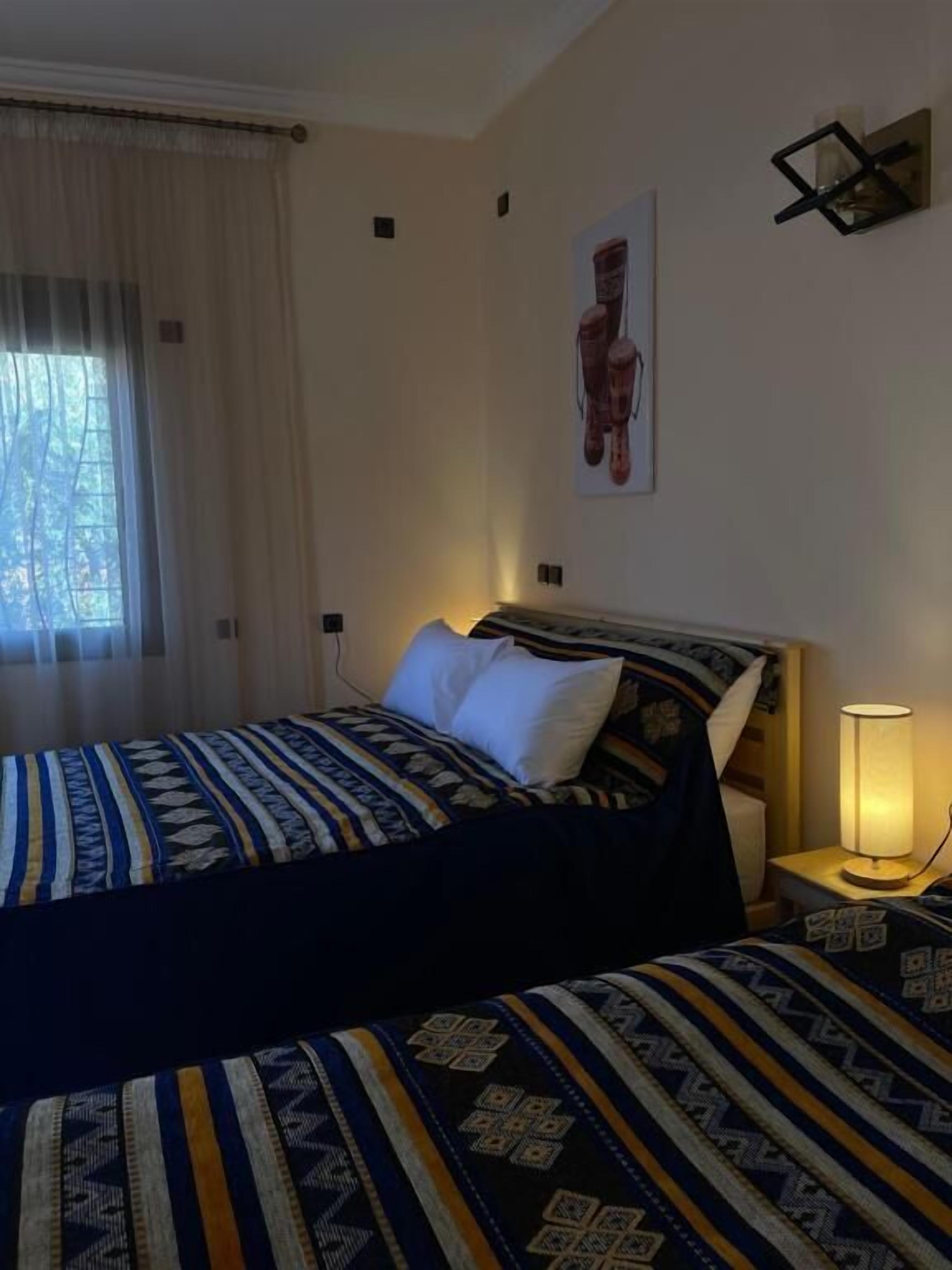 Une chambre d’hôtel comprenant un lit, une table de chevet avec une lampe, une fenêtre avec des rideaux et une œuvre d’art accrochée au mur.