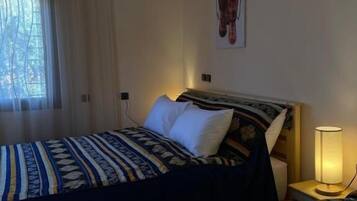 A hotel room with a bed, a nightstand with a lamp, a window with curtains, and a wall-mounted artwork.