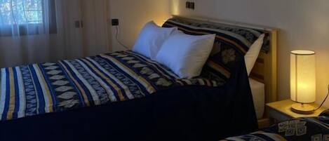 Une chambre d’hôtel comprenant un lit, une table de chevet avec une lampe, une fenêtre avec des rideaux et une œuvre d’art accrochée au mur.