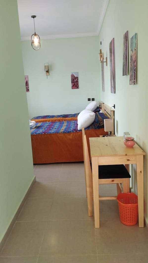 Una habitación con una cama, una mesa de madera, una silla, una lámpara colgante y un estante en la pared con objetos.