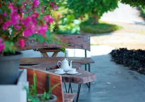 A wooden outdoor table with a white teapot and cups, surrounded by greenery and flowers.