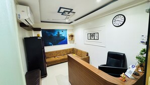 A reception area with a wooden desk, black office chair, and a sofa. There is a clock on the wall and a framed picture above the sofa. - Hotel Palladium (Surat)