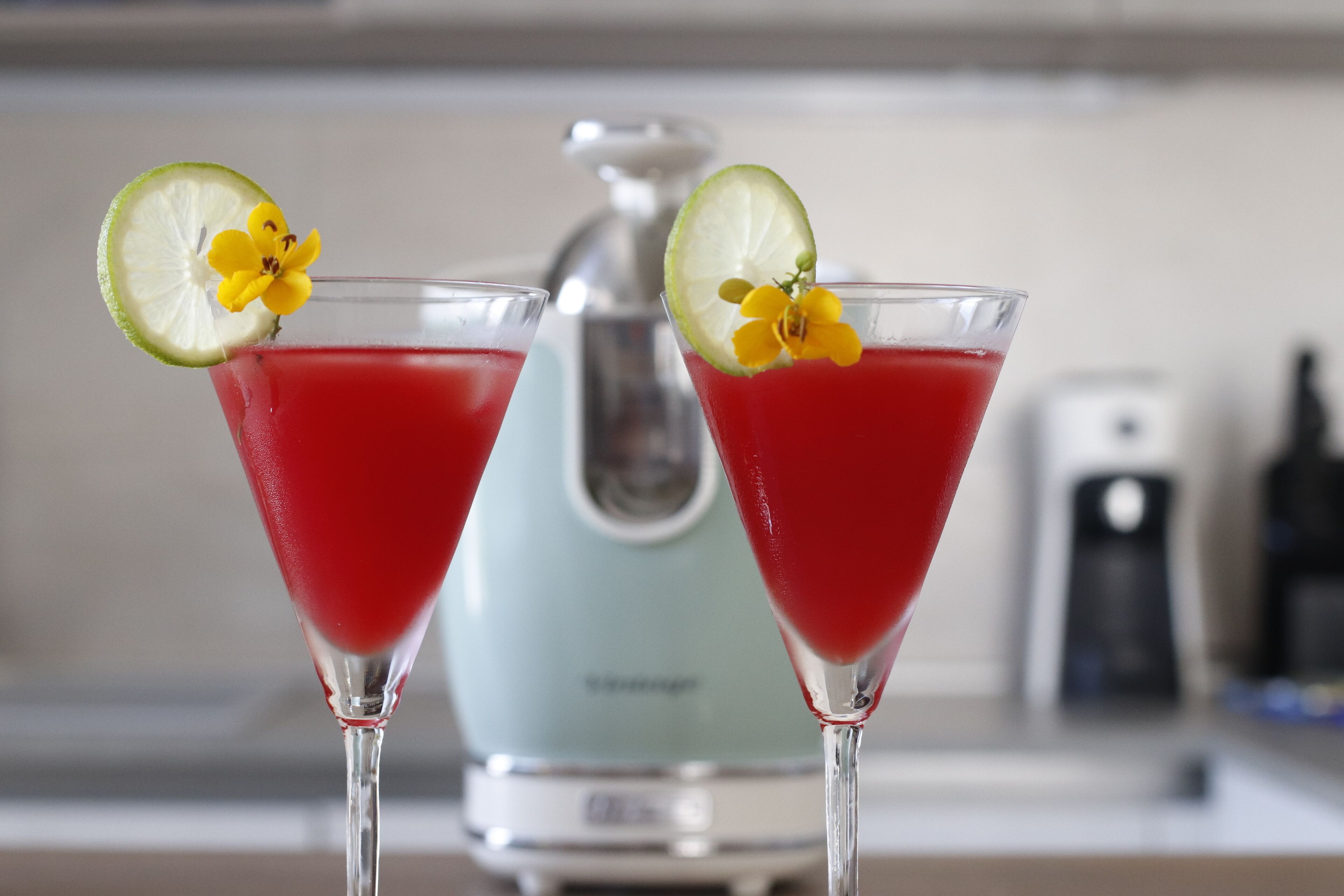 Two martini glasses with red liquid and lime slices, garnished with yellow flowers, in a kitchen setting with a blender in the background.