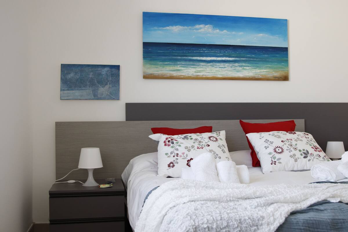 A neatly made bed with white linens and a blue ocean view painting above it.