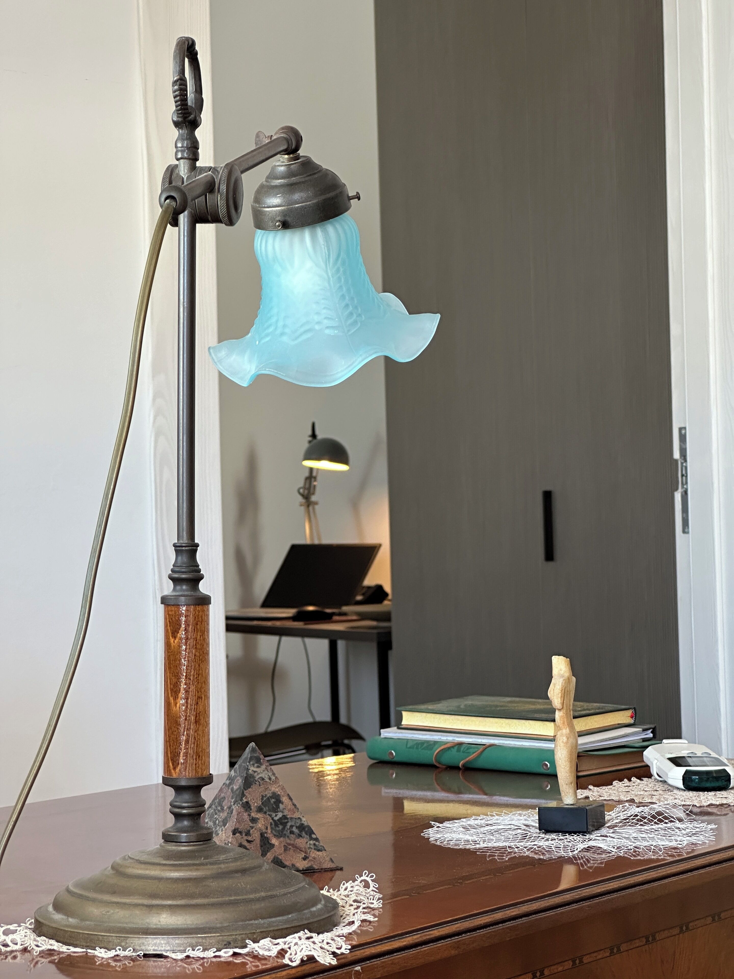 Una lámpara de escritorio con base de madera y pantalla de vidrio azul, una computadora portátil, libros y un pequeño objeto de madera sobre el escritorio.