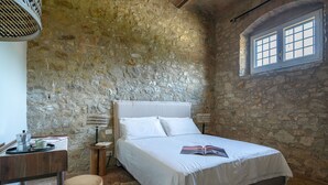 Una camera da letto con parete in pietra, un letto con biancheria bianca, una finestra a griglia e un tavolino con una lampada.