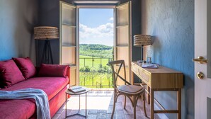Una habitación acogedora con un sofá rojo, una pequeña mesa redonda con una silla, y un escritorio con una silla. Hay dos lámparas a ambos lados de la puerta, que da a un balcón con vistas a un paisaje verde.