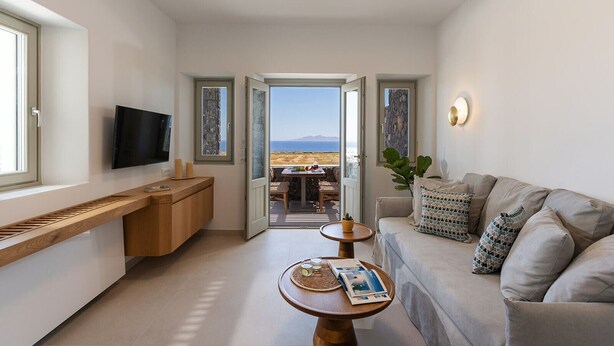 A modern, well-lit room with a TV, a wooden bench, a sofa with patterned cushions, and a view of the sea through an open door.