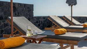 A rooftop lounge area with stone walls, wooden lounge chairs, and a table with a glass of water and a bottle.