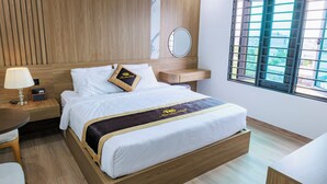 Habitación de hotel con una cama de madera, una mesita de noche con lámpara, un espejo redondo pequeño y una ventana con vistas a edificios.